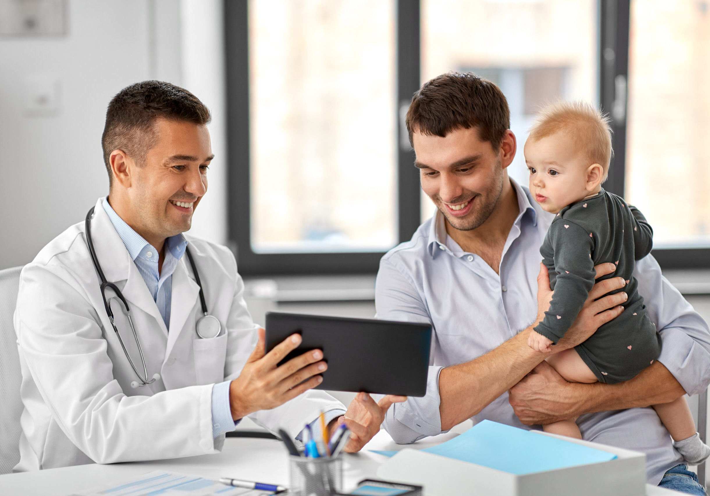 medicine, healthcare, pediatry and people concept - happy doctor showing tablet computer to father with baby daughter at medical office in hospital